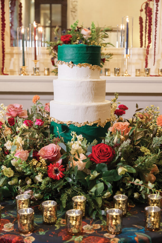 Three-tier green and white cake with gold accents, flowers, and gold candles on a decorative table in an elegant setting.