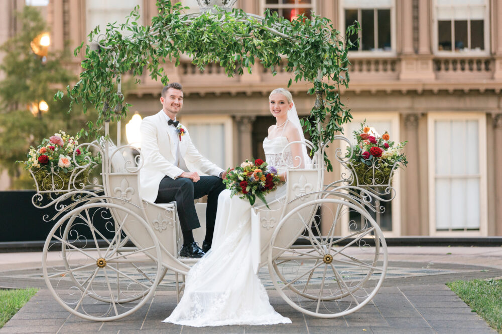 Bride and groom in formalwear smile on a white carriage with flowers, outdoors by a large, elegant building.