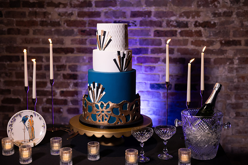 Three-tier cake with blue, white, and black geometric design, lit candles, crystal glasses, and bottle against a brick wall.