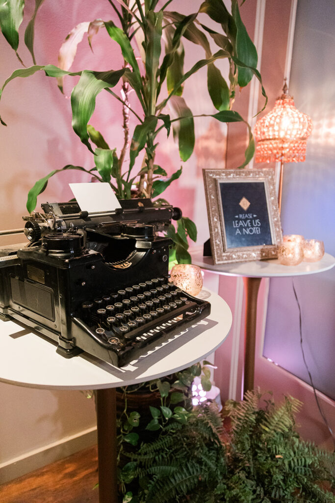 Vintage typewriter on a round table with a leafy plant, sign: “Please leave us a note!” Cozy vibe from candles and soft lighting.