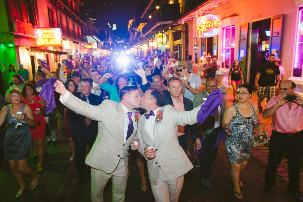 Two grooms kiss and hold purple cloths, leading a joyful crowd down a neon-lit street at night with friends and onlookers.