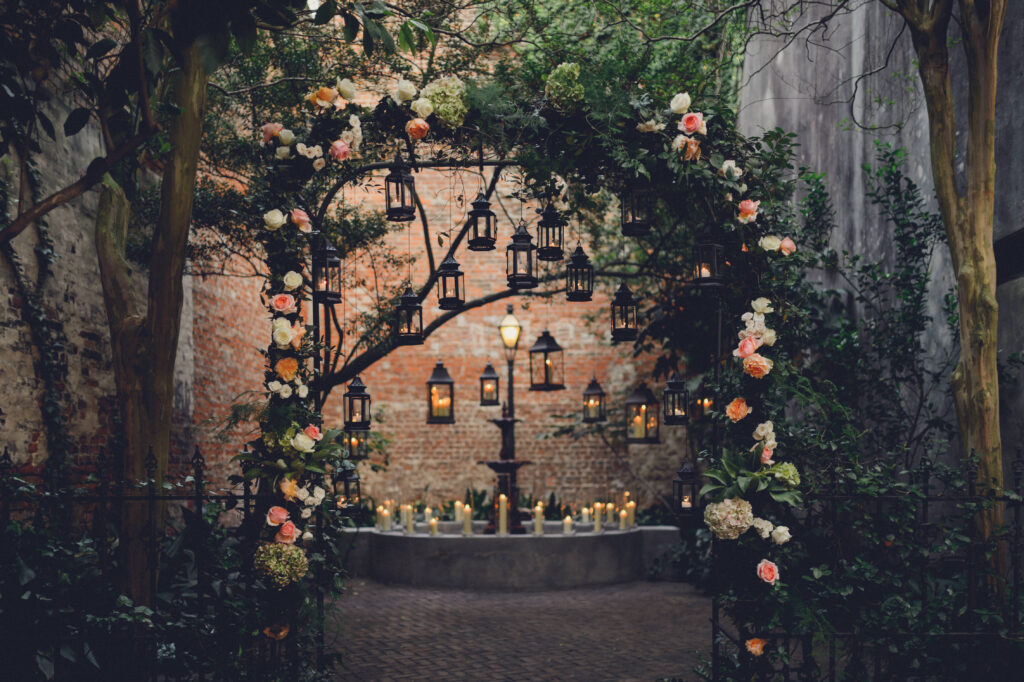 Floral arch with lanterns on a cobblestone path, brick wall, fountain, greenery, and candles create a romantic outdoor scene.