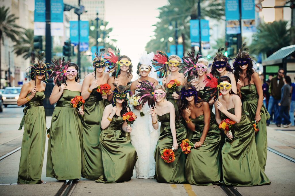 Bride in white and bridesmaids in green with feathered masks and bouquets pose on a city street.