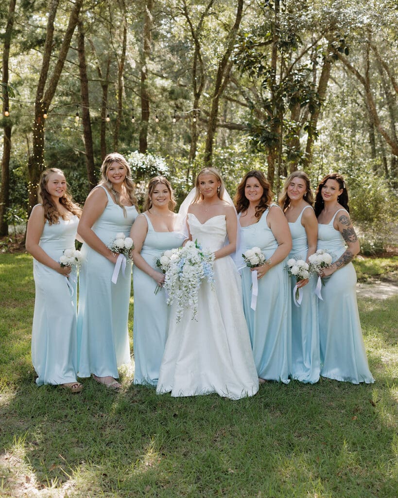 Bride in white gown with six bridesmaids in light blue dresses, holding bouquets outdoors among sunlit trees.