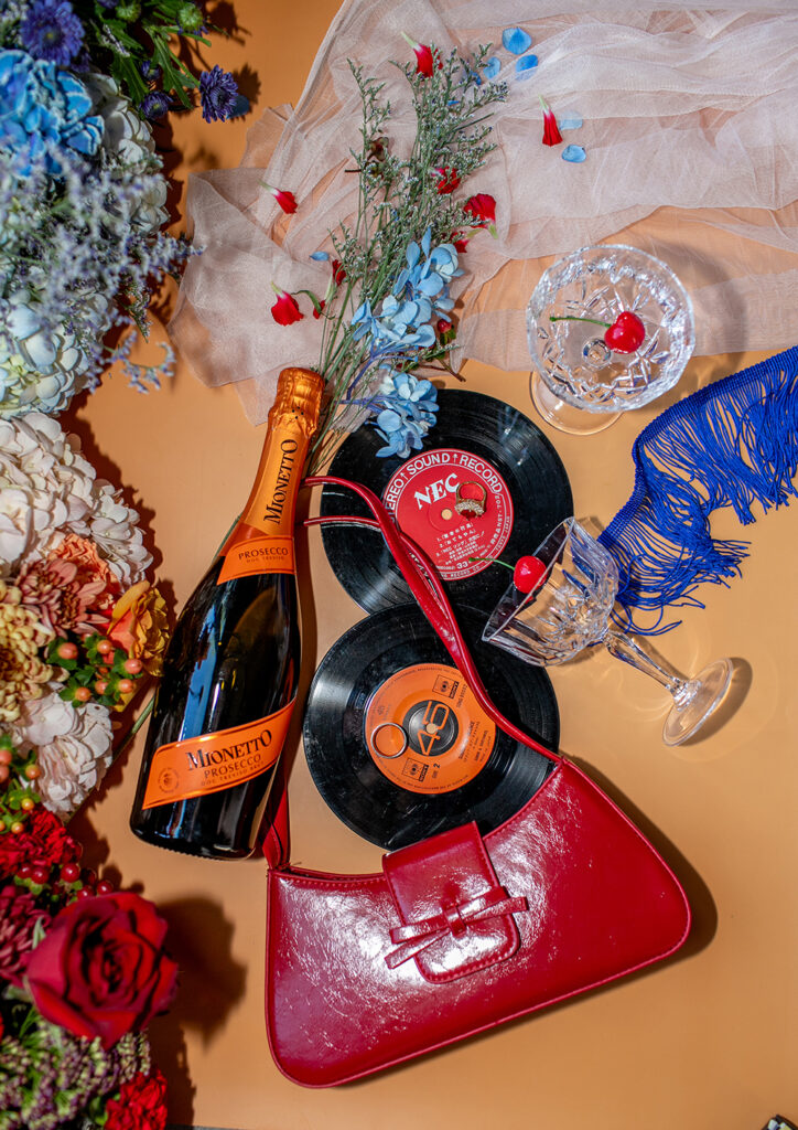 Mionetto prosecco, red handbag, vinyl records, cherry glass, flowers, and blue scarf on peach surface with sheer fabric.