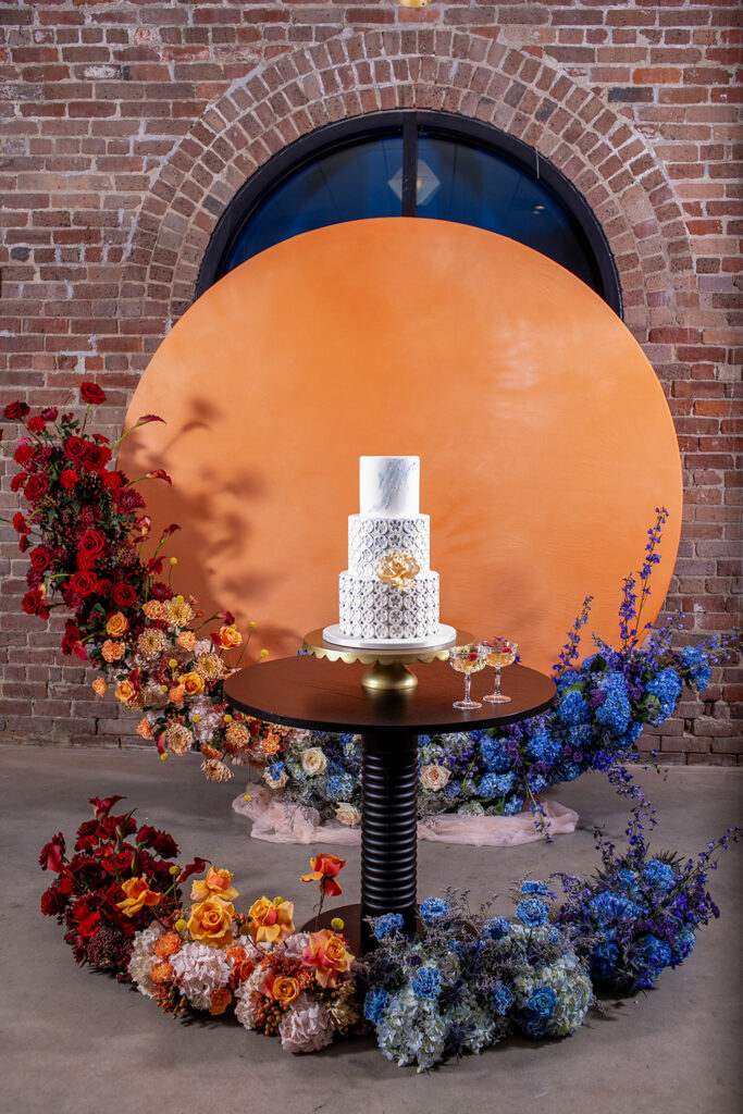 Three-tiered white cake on a round table with red, orange, and blue flowers, orange backdrop, and brick wall with arched window.