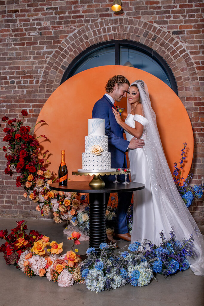 Bride and groom smile by a decorated cake table with colorful flowers, brick wall, and large orange circle backdrop.