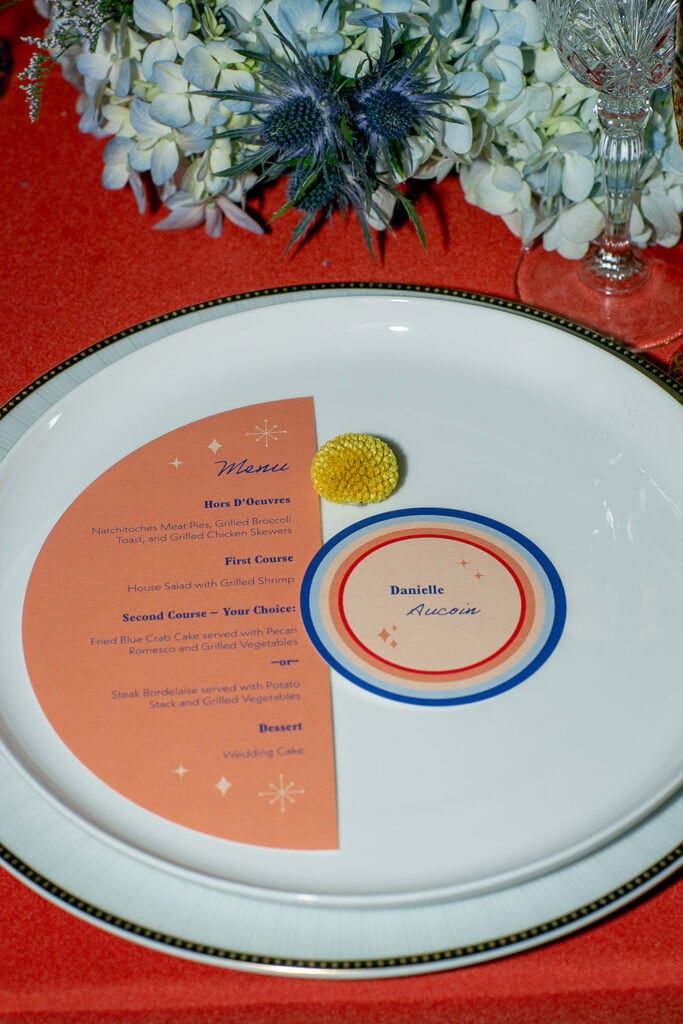 Decorative dinner plate with a colorful menu and Danielle Aucoin place card, next to a yellow flower and glassware.