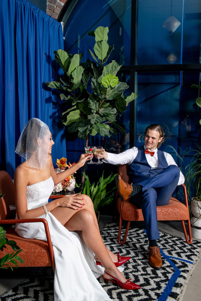 Bride in strapless dress and groom in blue suit toast drinks indoors, smiling with plants and blue curtains behind them.