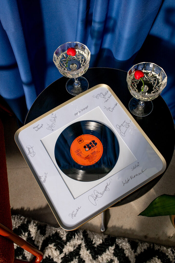 Signed vinyl record in frame on black table with two cherry-topped crystal glasses, blue curtains, and patterned rug behind.