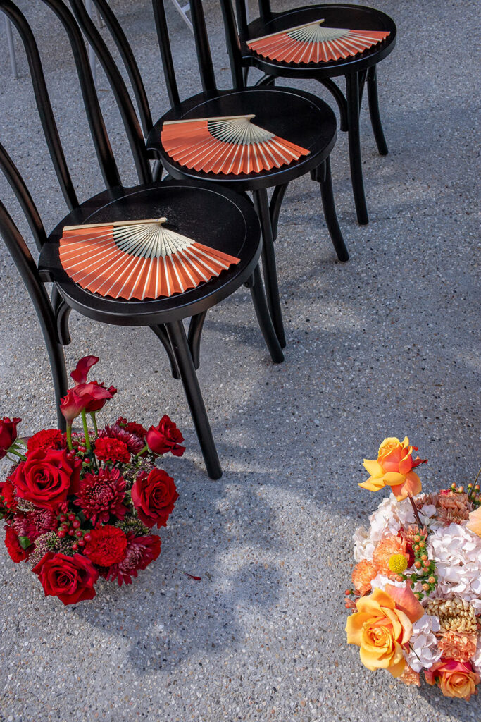 Three black chairs with orange fans are lined up, with two floral arrangements—red and multicolored—on the ground nearby.