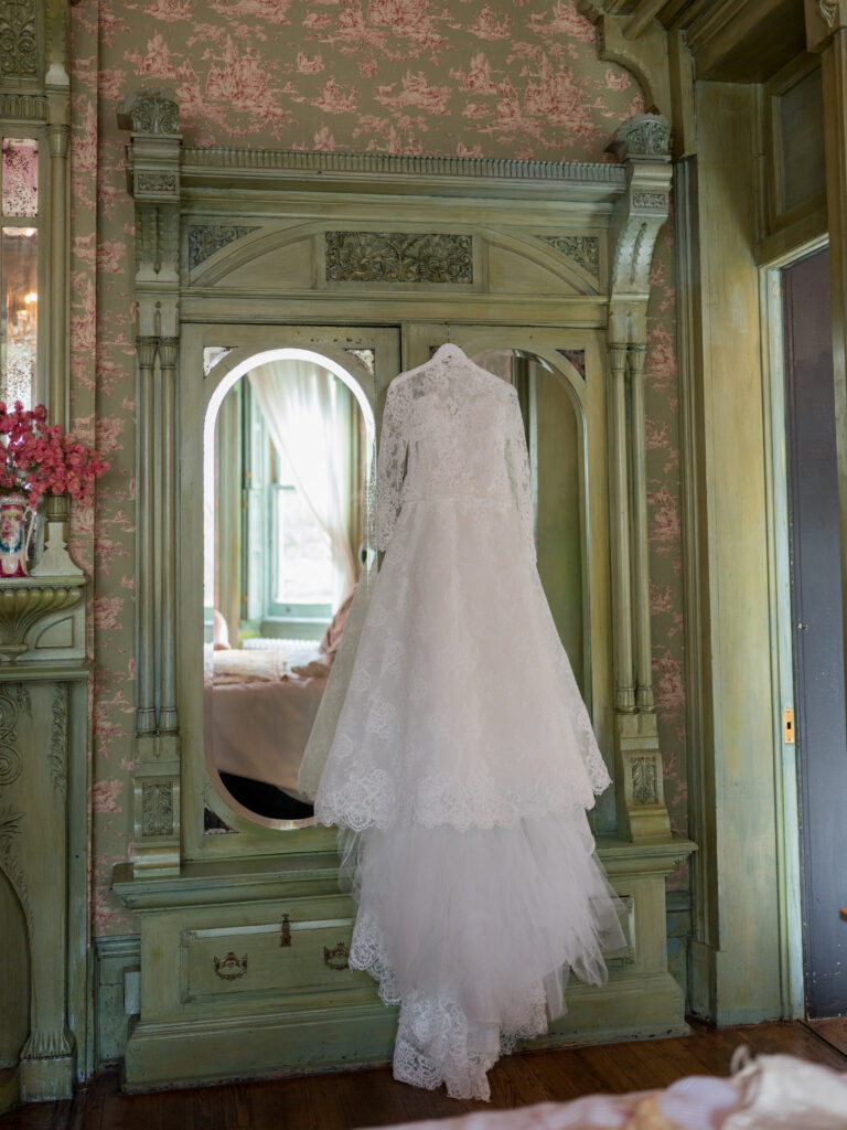 Long-sleeve lace wedding dress hangs from an antique mirror inside a vintage room with ornate green woodwork and toile-patterned wallpaper. Soft natural light filters through the windows, illuminating the gown’s intricate lace details and layered tulle skirt, creating a romantic and timeless bridal detail shot.
