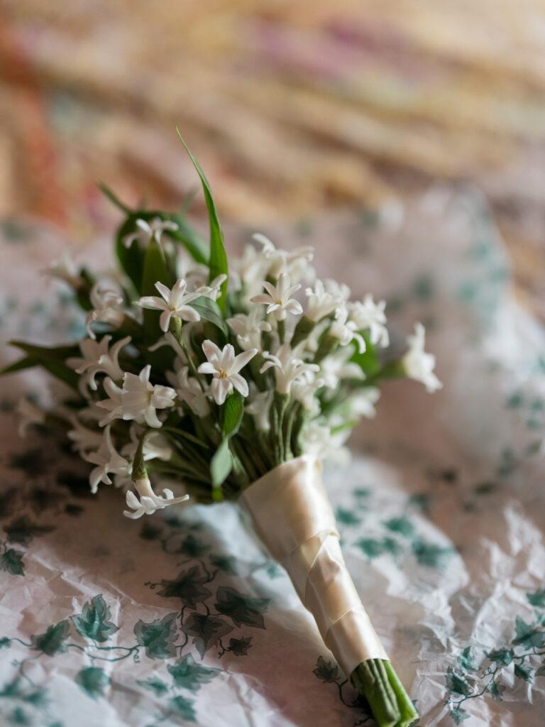Close-up of a small bridal bouquet made of delicate white flowers and green leaves, wrapped with ivory ribbon. The bouquet rests on floral-patterned tissue paper with soft, warm light creating a romantic and intimate wedding detail shot.
