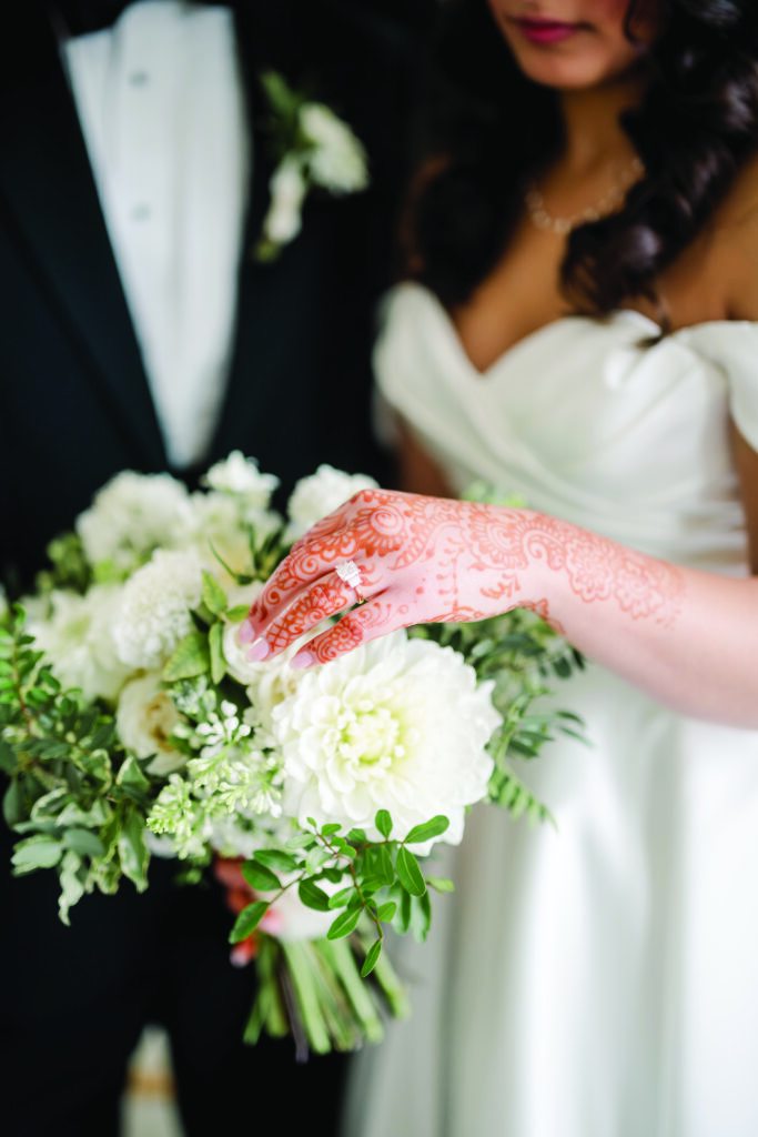 A bride’s henna-adorned hand, wearing her engagement ring, gently rests on a bouquet of white dahlias and greenery. Part of her off-the-shoulder white gown and the groom’s black tuxedo are softly visible in the background.