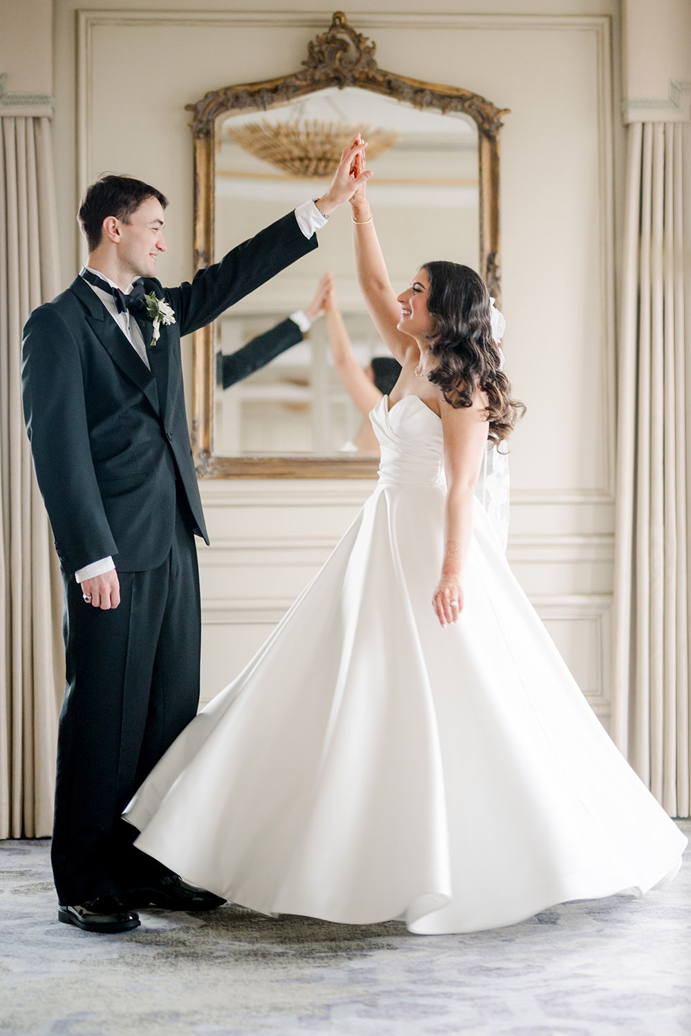 A bride and groom dance joyfully in an elegant room, with the bride twirling in her strapless white gown and veil, and the groom in a black tuxedo holding her hand. Behind them, a large ornate mirror and chandelier add to the romantic, formal atmosphere.