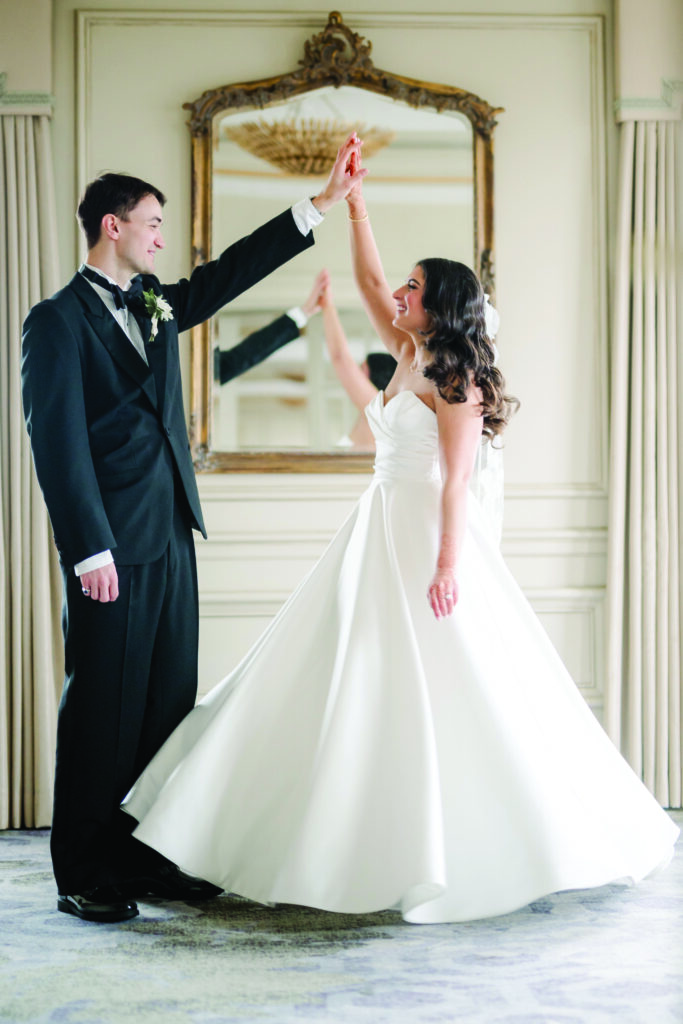 A bride and groom dance joyfully in an elegant room, with the bride twirling in her strapless white gown and veil, and the groom in a black tuxedo holding her hand. Behind them, a large ornate mirror and chandelier add to the romantic, formal atmosphere.