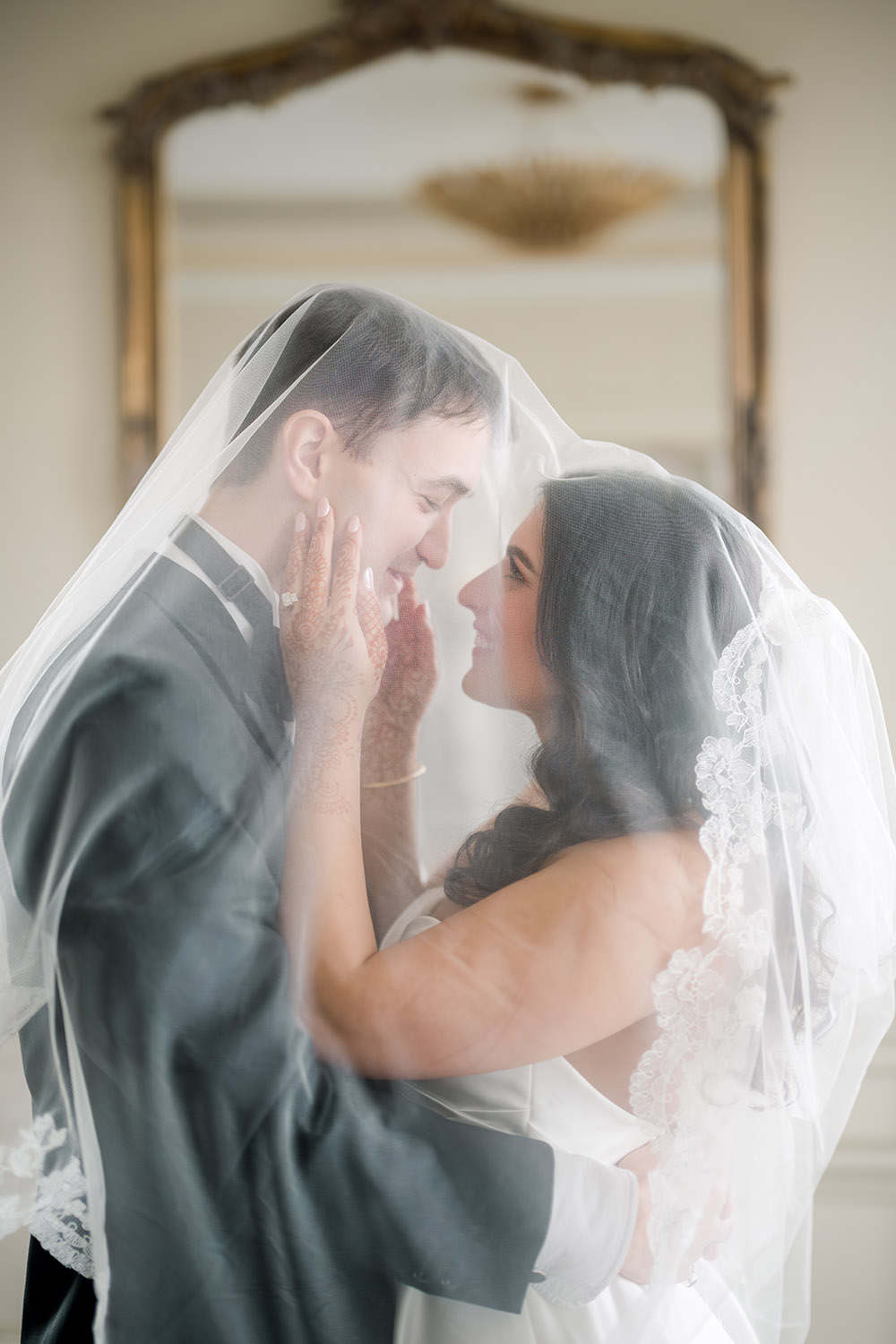 Bride and groom stand forehead to forehead beneath a delicate lace-edged veil, smiling softly as she cups his face with hands adorned in henna. The groom holds her waist as they share an intimate, romantic moment in a softly lit room with an ornate mirror in the background.