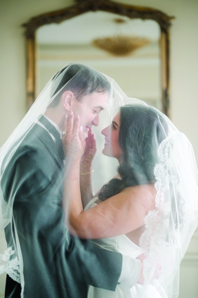 Bride and groom stand forehead to forehead beneath a delicate lace-edged veil, smiling softly as she cups his face with hands adorned in henna. The groom holds her waist as they share an intimate, romantic moment in a softly lit room with an ornate mirror in the background.