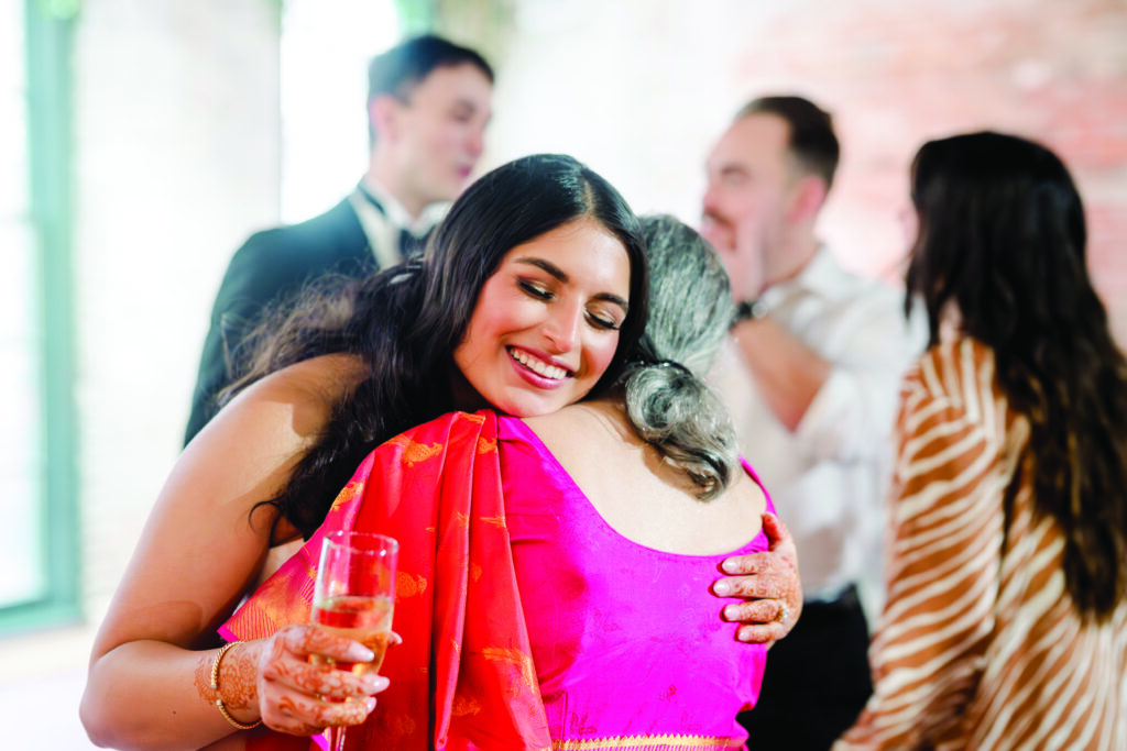“A smiling woman in a bright red and gold sari hugs an older woman during a celebration, holding a champagne glass in one hand. The moment is warm and joyful, with other guests chatting in the softly blurred background.”