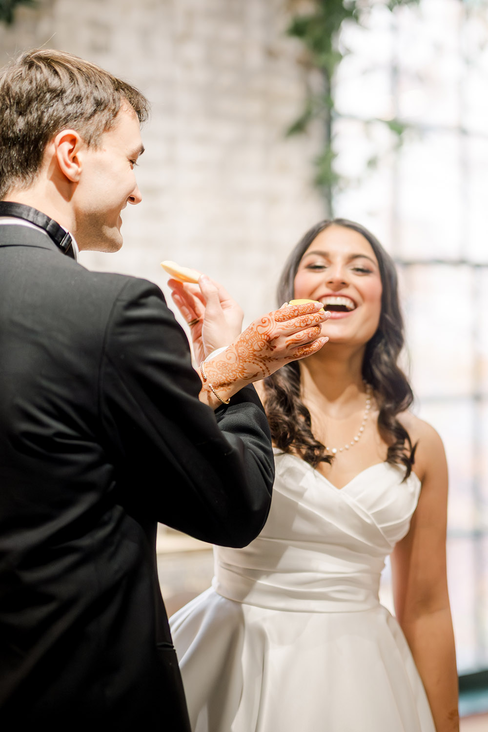 Bride and groom stand forehead to forehead beneath a delicate lace-edged veil, smiling softly as she cups his face with hands adorned in henna. The groom holds her waist as they share an intimate, romantic moment in a softly lit room with an ornate mirror in the background.