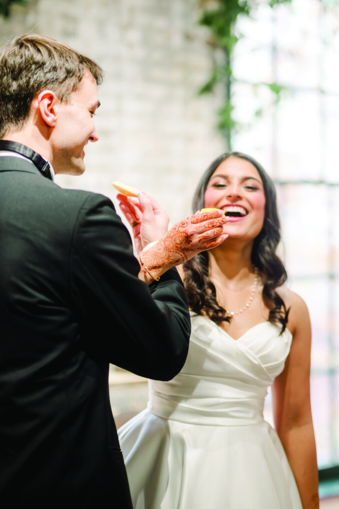 A groom in a black tuxedo feeds a cookie to his smiling bride, who wears a white gown and pearl necklace. Her henna-decorated hand holds another cookie as they share a playful moment during their wedding reception.