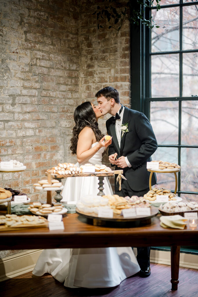 A bride and groom share a sweet kiss in front of a dessert table filled with cookies and pastries. The bride in a white gown holds a lemon bar while the groom in a black tuxedo leans in, standing against a rustic brick wall and large window.