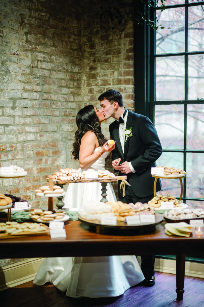 “A bride and groom share a sweet kiss in front of a dessert table filled with cookies and pastries. The bride in a white gown holds a lemon bar while the groom in a black tuxedo leans in, standing against a rustic brick wall and large window.”