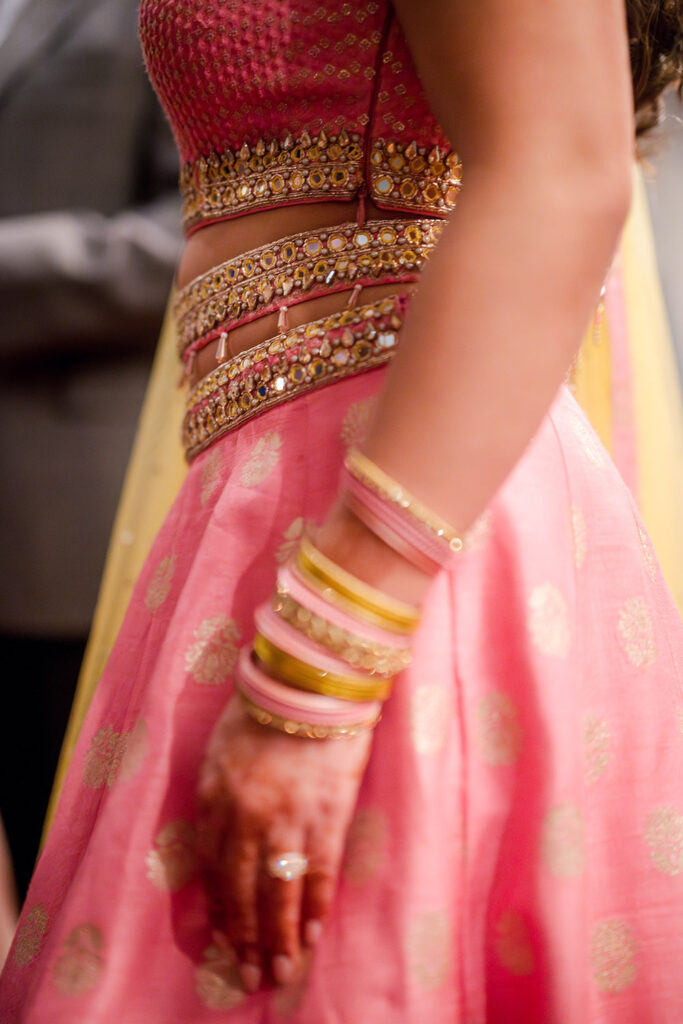 Close-up of a woman wearing a pink and gold embellished lehenga with mirrorwork details, henna on her hand, and stacked pink and gold bangles as she stands at an Indian pre-wedding celebration