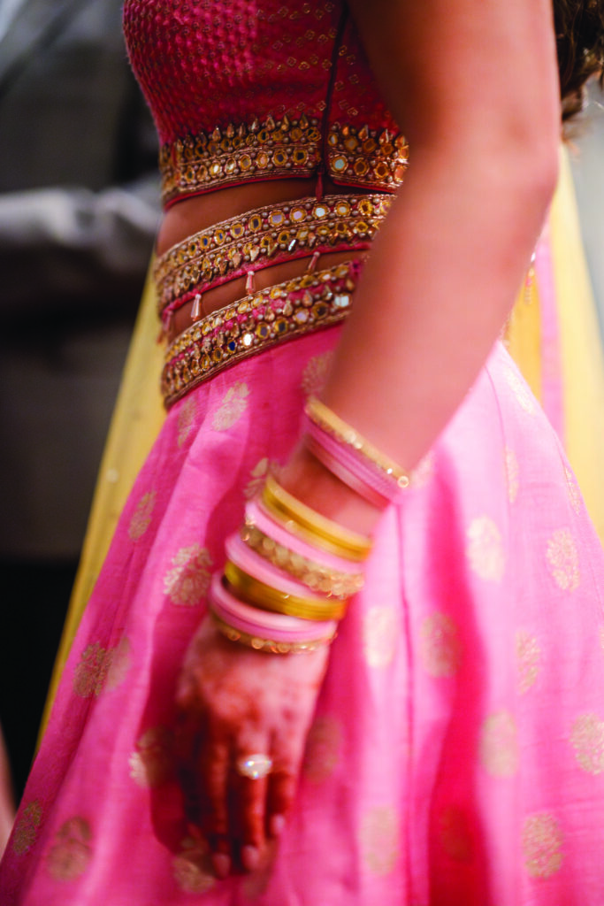 Here’s a clear, elegant alt text for this image:

**Alt Text:**
“Close-up of a woman wearing a pink and gold embellished lehenga with mirrorwork details, henna on her hand, and stacked pink and gold bangles as she stands at an Indian pre-wedding celebration.”
