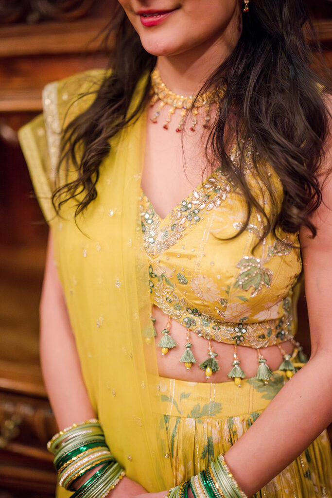 Close-up of a woman wearing a yellow embellished lehenga with floral embroidery, green tassel details, and matching green and gold bangles, paired with a gold necklace and soft waves in her hair