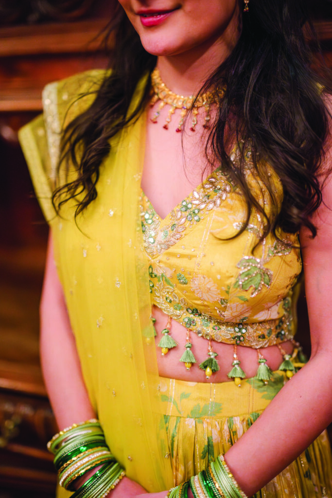 Here’s a clear, elegant, and descriptive alt text for this image:

**Alt Text:**
“Close-up of a woman wearing a yellow embellished lehenga with floral embroidery, green tassel details, and matching green and gold bangles, paired with a gold necklace and soft waves in her hair.”
