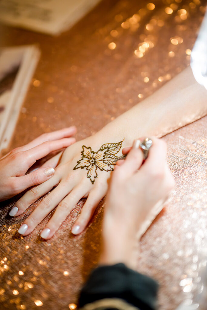 Close-up of a mehndi artist applying henna in a floral design on the back of a woman’s hand, with intricate lines forming petals and leaves over a sparkling sequin tablecloth at an Indian pre-wedding event.