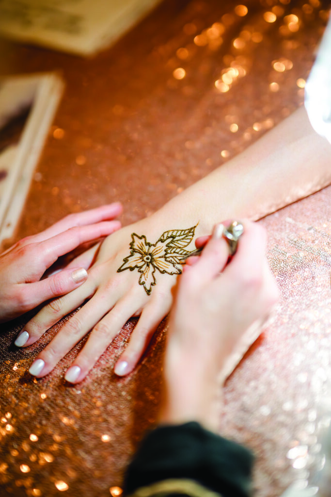 Close-up of a mehndi artist applying henna in a floral design on the back of a woman’s hand, with intricate lines forming petals and leaves over a sparkling sequin tablecloth at an Indian pre-wedding event.