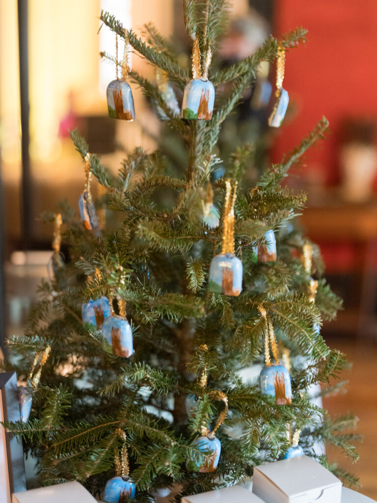 A small evergreen Christmas tree decorated with hand-painted blue and brown ornaments, each hanging from a gold ribbon. The tree sits indoors with soft, warm lighting in the background, surrounded by white gift boxes at its base.