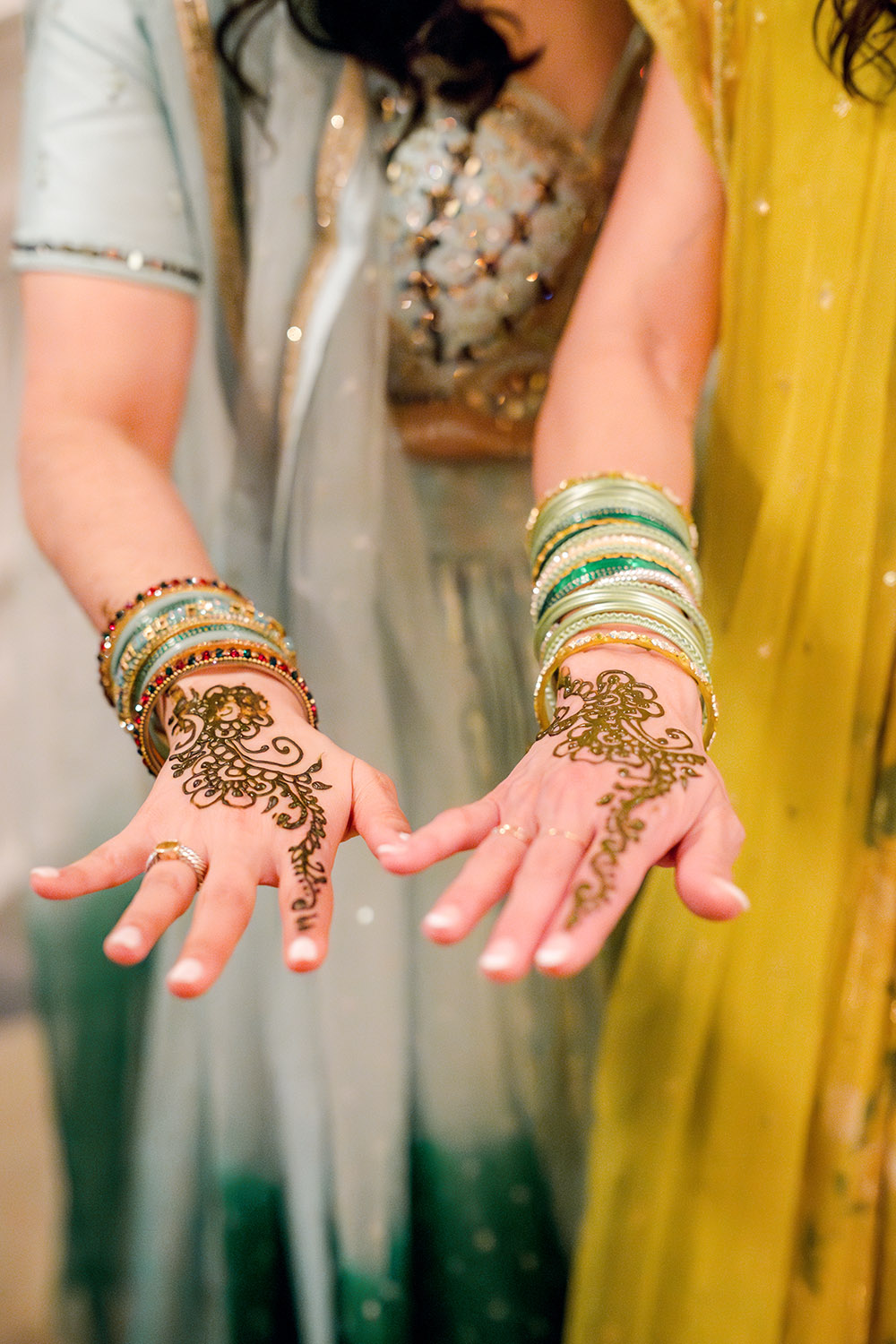 Two women extend their hands to show fresh mehndi designs on their palms, accented by colorful bangles. One wears a light blue lehenga and the other a yellow outfit, highlighting the intricate henna patterns.