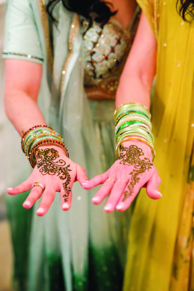 Here’s a clear, descriptive, and culturally accurate alt text for this image:

**Alt Text:**
“Two women extend their hands to show fresh mehndi designs on their palms, accented by colorful bangles. One wears a light blue lehenga and the other a yellow outfit, highlighting the intricate henna patterns.”

