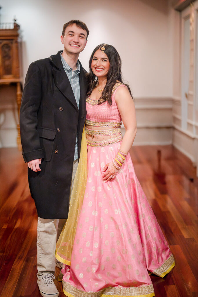 Couple smiling together at an Indian pre-wedding celebration; woman wearing a pink and gold lehenga with henna on her hands and traditional jewelry, standing beside her partner in a dark coat inside a warmly lit venue with polished wood floors.