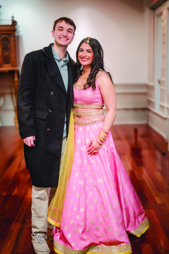 Couple smiling together at an Indian pre-wedding celebration; woman wearing a pink and gold lehenga with henna on her hands and traditional jewelry, standing beside her partner in a dark coat inside a warmly lit venue with polished wood floors.