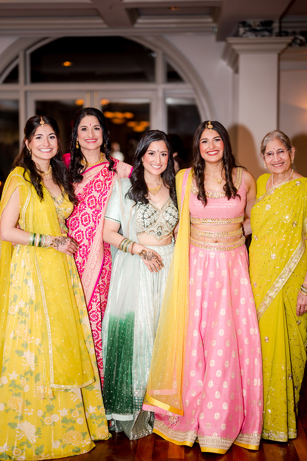 Group of women dressed in vibrant Indian attire at a pre-wedding celebration, wearing colorful lehengas and saris in yellow, pink, mint green, and red. They stand smiling together, showcasing traditional jewelry, henna designs, and elegant embroidery in a warm indoor venue.