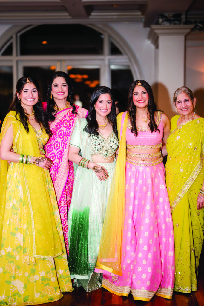 Group of women dressed in vibrant Indian attire at a pre-wedding celebration, wearing colorful lehengas and saris in yellow, pink, mint green, and red. They stand smiling together, showcasing traditional jewelry, henna designs, and elegant embroidery in a warm indoor venue.