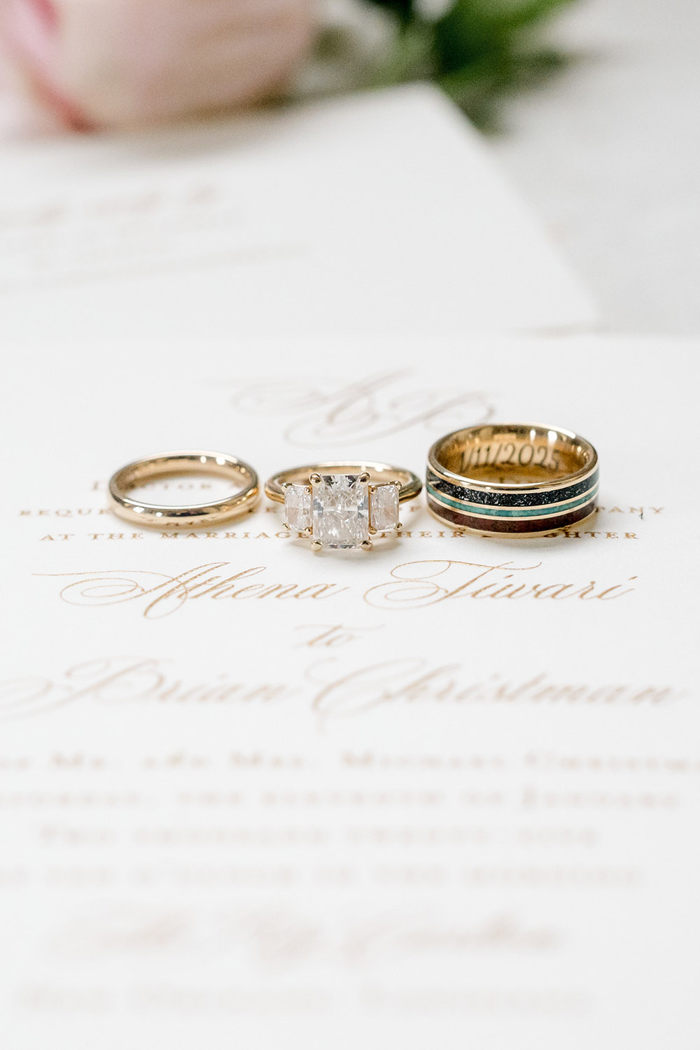 Close-up of three wedding rings displayed on top of an elegant ivory invitation with gold script lettering. The set includes a simple gold band, a three-stone princess-cut diamond engagement ring, and a gold men’s band with dark inlay and engraved date. Soft florals blur in the background.