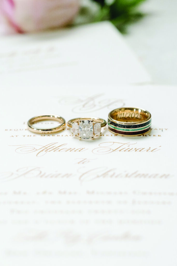 Close-up of three wedding rings displayed on top of an elegant ivory invitation with gold script lettering. The set includes a simple gold band, a three-stone princess-cut diamond engagement ring, and a gold men’s band with dark inlay and engraved date. Soft florals blur in the background.