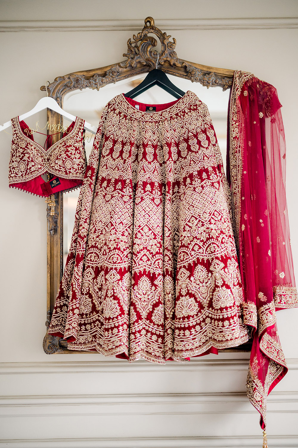 Red and gold embroidered bridal lehenga displayed on ornate hangers, including the heavily detailed skirt, matching embellished blouse, and sheer dupatta with gold trim, all hanging against an antique gold-framed mirror.