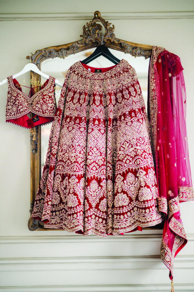 Red and gold embroidered bridal lehenga displayed on ornate hangers, including the heavily detailed skirt, matching embellished blouse, and sheer dupatta with gold trim, all hanging against an antique gold-framed mirror.