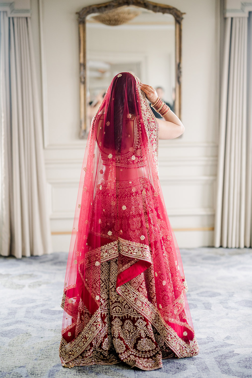 Bride seen from behind adjusting her red and gold embroidered dupatta, wearing a traditional lehenga with intricate beadwork and patterns. She stands in an elegant room with soft natural light, facing an ornate mirror and flowing curtains.