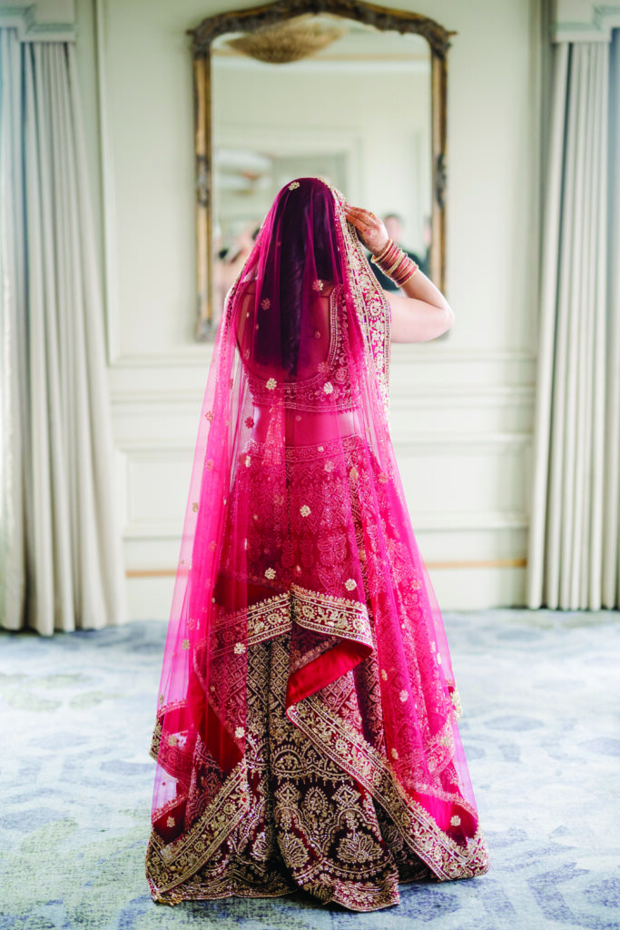 Bride seen from behind adjusting her red and gold embroidered dupatta, wearing a traditional lehenga with intricate beadwork and patterns. She stands in an elegant room with soft natural light, facing an ornate mirror and flowing curtains.