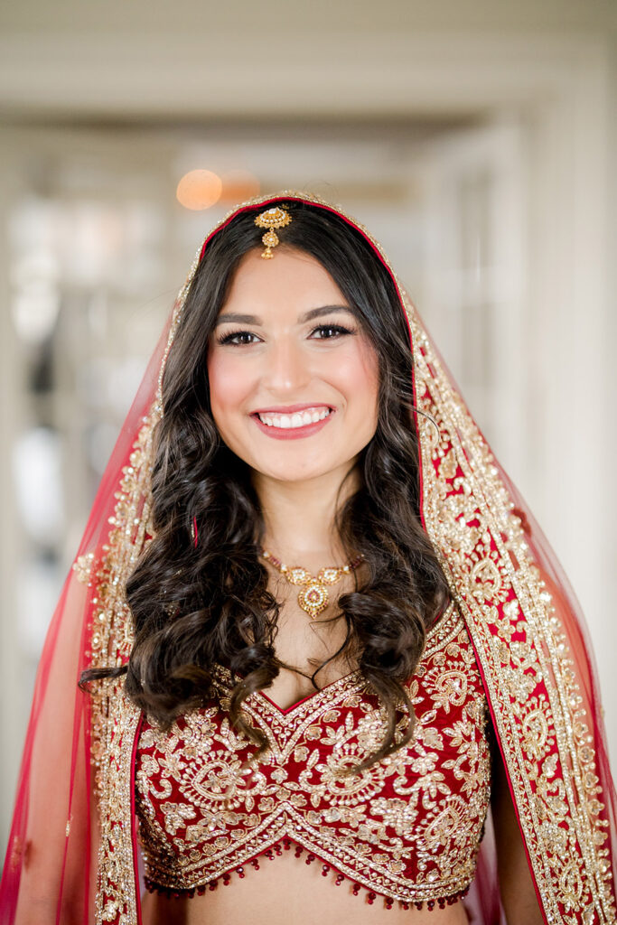 Bride smiling brightly while wearing a traditional red and gold embroidered lehenga with a matching dupatta draped over her head, featuring intricate beadwork and floral patterns. She has soft curls, elegant bridal jewelry, and a gold maang tikka, standing in soft indoor light.