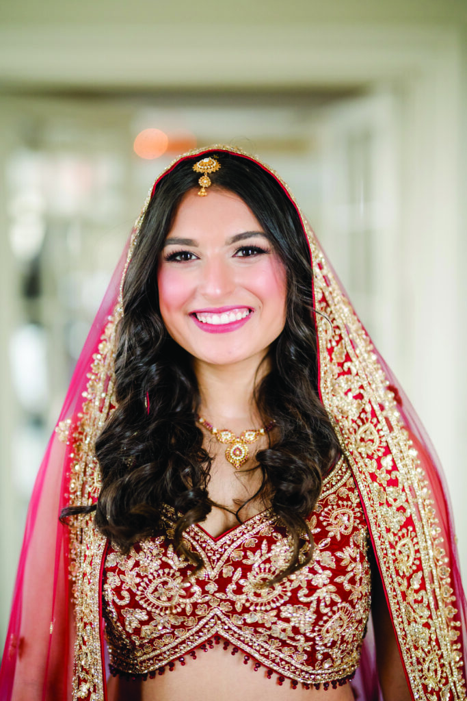 Bride smiling brightly while wearing a traditional red and gold embroidered lehenga with a matching dupatta draped over her head, featuring intricate beadwork and floral patterns. She has soft curls, elegant bridal jewelry, and a gold maang tikka, standing in soft indoor light.