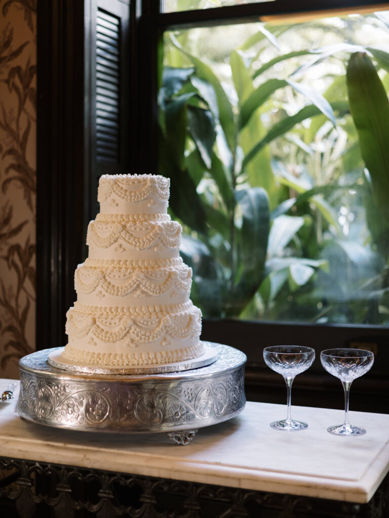 A four-tier white wedding cake decorated with intricate piped lace and pearl details sits on an ornate silver cake stand. Two crystal coupe glasses rest beside it on a marble table, with lush green foliage visible through the window behind.