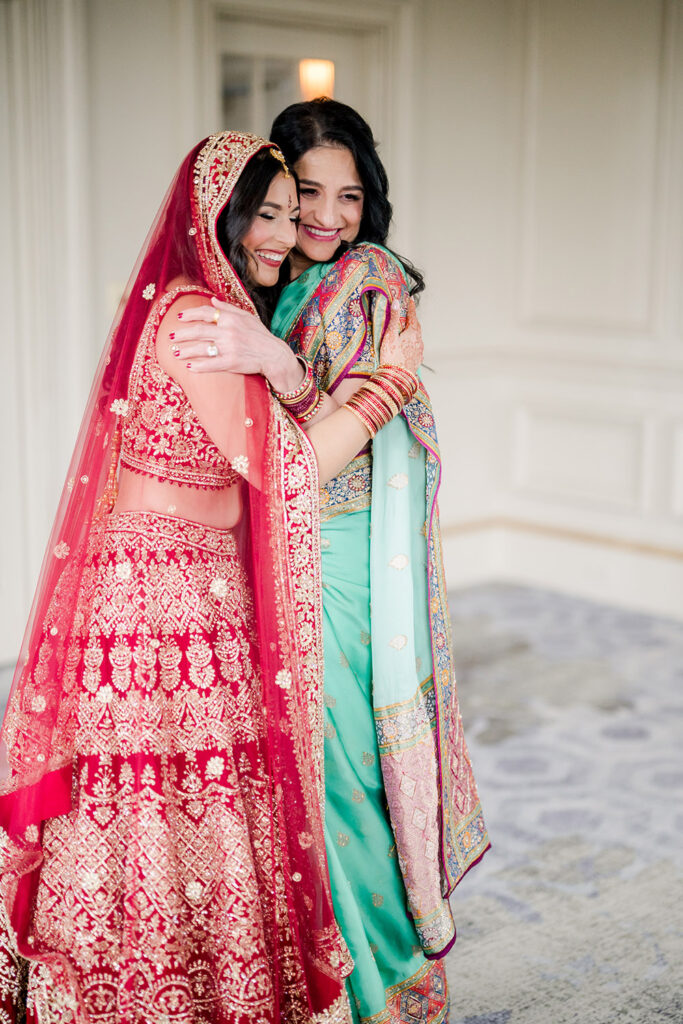 Bride in a red and gold embroidered lehenga warmly embraces a woman wearing a teal saree with colorful embroidered details. Both smile with eyes closed, sharing an emotional moment indoors before the ceremony.